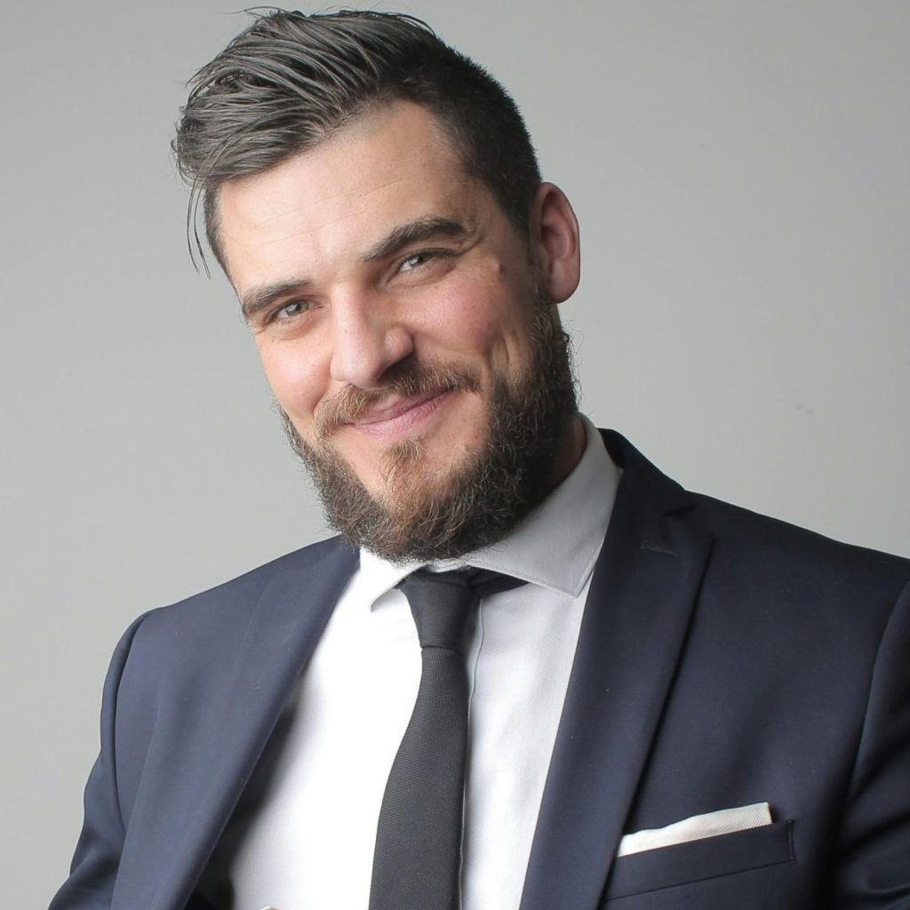 Smiling bearded man in a suit and tie with a pocket square against a grey background