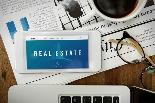 Tablet displaying real estate website on desk with newspaper, glasses, coffee, laptop keyboard nearby